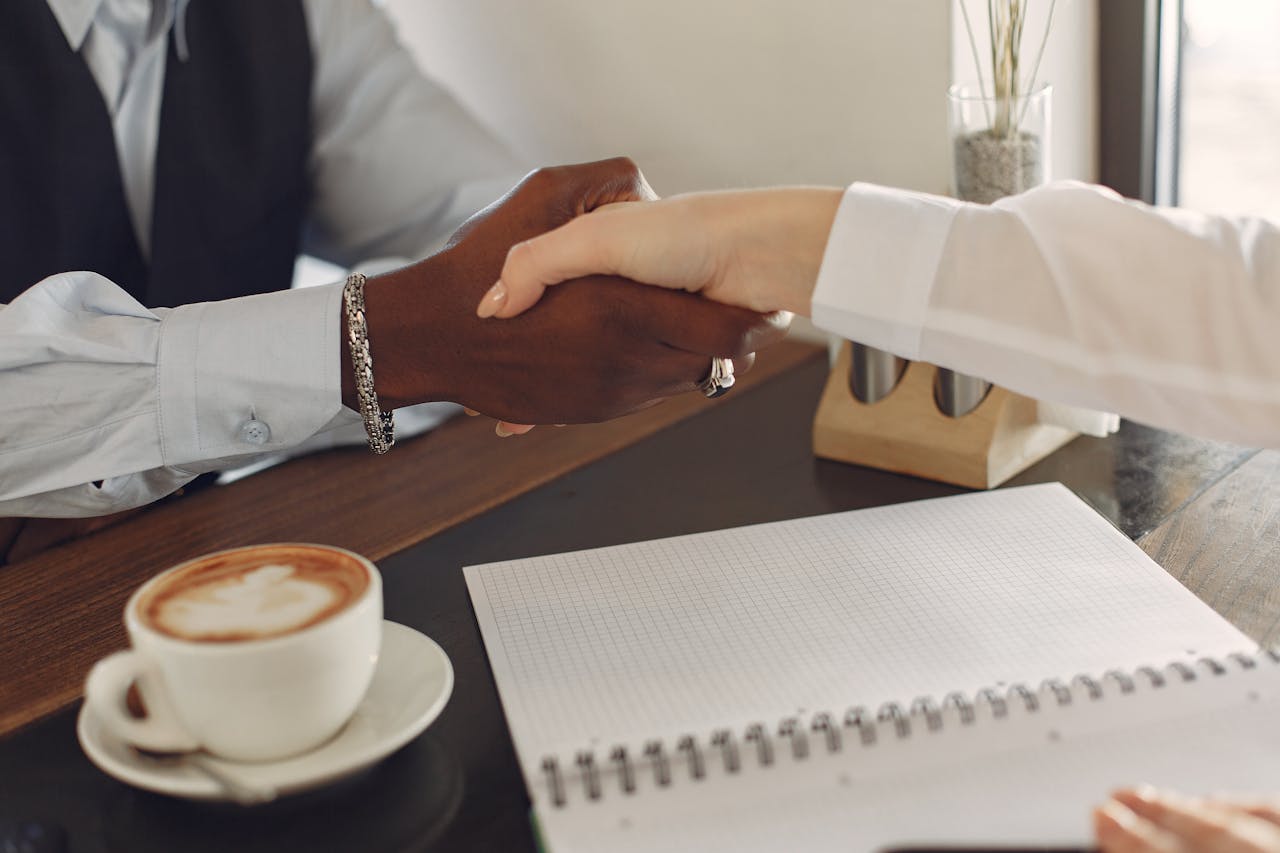 gallery-6 Two professionals shaking hands at a desk with a coffee cup and notebook, symbolizing business agreement.